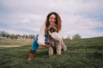 A beautiful woman is in the meadow with her dog. The owner is hugging her pet while trying to take a photo with her phone. The pet is a Spanish water dog with brown fur.