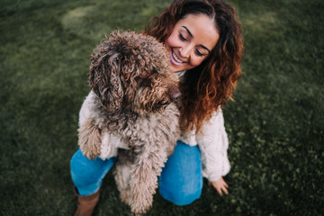 A beautiful woman is in the meadow with her dog. The owner is hugging her pet while looking at it with love. They are enjoying a day in the park. The pet is a Spanish water dog with brown fur.