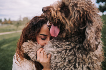A beautiful woman is in the meadow with her dog. They are hugging. She has her eyes closed while the doggy is looking far. They are enjoying their company. The pet is a Spanish water dog.