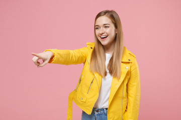 Cheerful young woman girl in yellow leather jacket posing isolated on pastel pink wall background studio portrait. People emotions lifestyle concept. Mock up copy space. Pointing index finger aside.