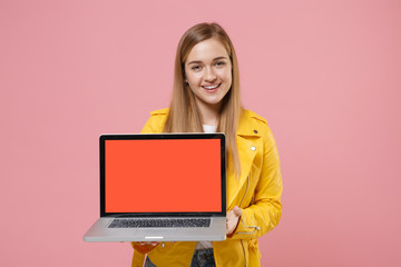 Naklejka premium Smiling young woman girl in yellow leather jacket posing isolated on pastel pink background in studio. People lifestyle concept. Mock up copy space. Hold laptop pc computer with blank empty screen.