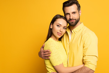 Handsome man embracing beautiful smiling girlfriend and looking at camera on yellow background