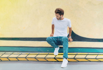 Young handsome guy is standing on a graffiti wall background. Man is wearing white, empty basic t-shirt without logo. Mock-up with light flare.