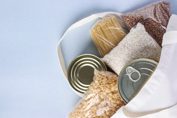 Textile Grocery Bag with food supplies crisis food stock for quarantine isolation period on light blue background. Rice, buckwheat, pasta, canned food. Food delivery, Donation, Top view