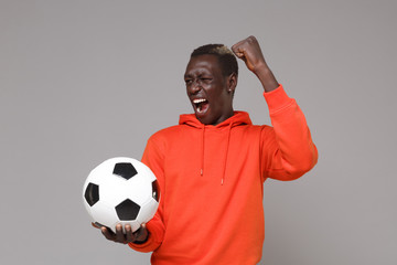 Fototapeta Screaming young african american man guy football player in orange streetwear hoodie isolated on grey background. Sport leisure lifestyle concept. Play football hold soccer ball doing winner gesture. obraz