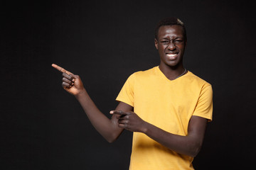 Cheerful young african american man guy in yellow t-shirt posing isolated on black wall background studio portrait. People emotions lifestyle concept. Mock up copy space. Pointing index fingers aside.