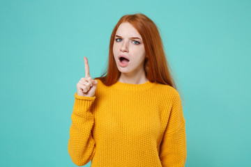 Dissatisfied shocked young redhead woman girl in yellow sweater posing isolated on blue turquoise background in studio. People lifestyle concept. Mock up copy space. Pointing index finger up swearing.