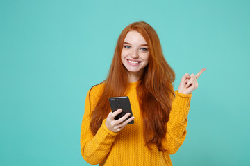 Smiling young redhead woman girl in yellow sweater posing isolated on blue turquoise background in studio. People lifestyle concept. Mock up copy space. Using mobile phone point index finger aside up.