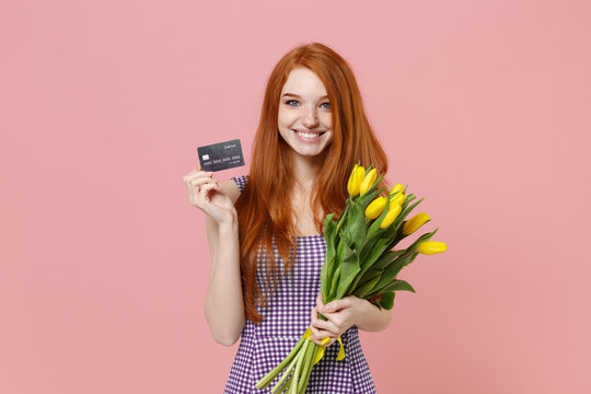 Smiling young redhead woman girl in plaid dress posing isolated on pastel pink wall background in studio. People lifestyle concept. Mock up copy space. Hold bouquet of yellow tulips, credit bank card. - Powered by Adobe