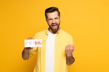 Cheerful man showing yeah gesture and holding gift card on yellow background