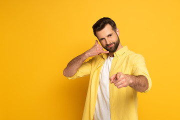 Fototapeta premium Handsome man showing call sign and pointing with finger at camera on yellow background