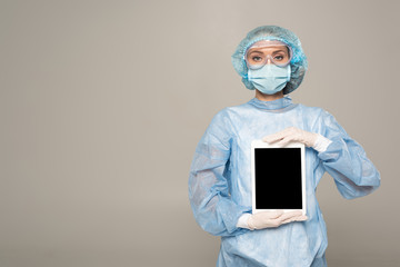 Doctor in safety googles and medical mask holding digital tablet with blank screen isolated on grey