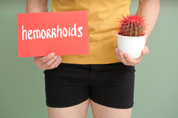 Young man holding cactus and paper with text HEMORRHOIDS on color background