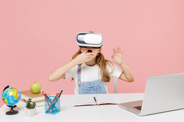 Shocked little kid schoolgirl 12-13 years old study at white desk with pc laptop isolated on pink background. School distance education at home concept. Watching in headset, covering mouth with hand.