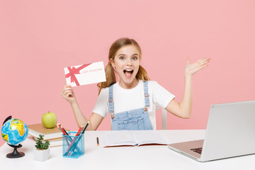 Amazed little kid schoolgirl 12-13 years old study at desk with laptop isolated on pink background. School distance education at home during quarantine concept. Hold gift certificate point hand aside.