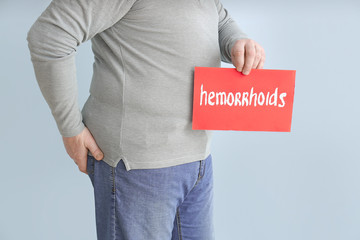Elderly man holding paper with text HEMORRHOIDS on color background