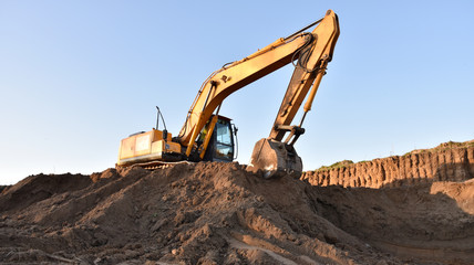 Fototapeta na wymiar Excavator working on earthmoving. Backhoe digs ground in sand quarry on blue sky background. Construction machinery for excavation, loading, lifting and hauling of cargo on job sites