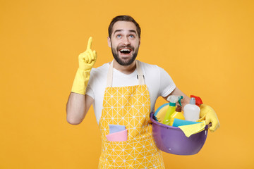 Excited young man househusband in apron rubber gloves hold basin with detergent bottles washing...