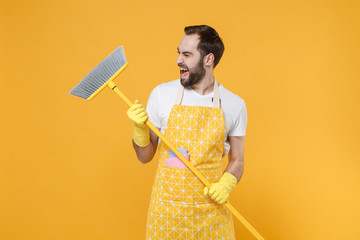 Laughing funny young man househusband in apron rubber gloves hold in hands broom while doing...