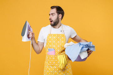 Perplexed young man househusband in apron hold basket with clean clothes, iron doing housework...