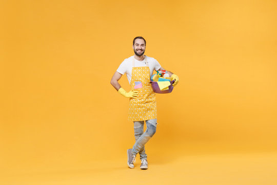 Smiling man househusband in apron rubber gloves hold basin detergent bottles washing cleansers doing housework isolated on yellow background. Housekeeping concept. Standing with arm akimbo on waist.