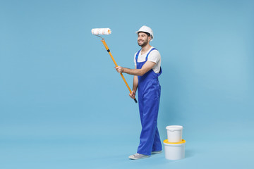 Funny young man in coveralls protective helmet hardhat hold paint roller, bucket isolated on pastel blue background studio. Instruments accessories for renovation apartment room. Repair home concept.