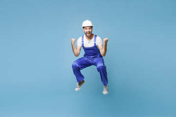 Joyful young man in coveralls protective helmet hardhat isolated on blue background studio. Instruments accessories for renovation apartment room. Repair home concept. Jumping, doing winner gesture.