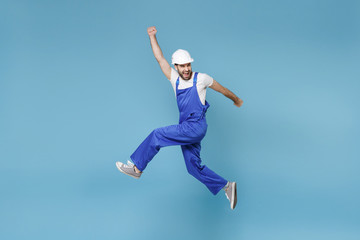 Fototapeta na wymiar Overjoyed young man in coveralls protective helmet hardhat isolated on blue background. Instruments accessories renovation apartment room. Repair home concept. Jumping clenching fists like winner.