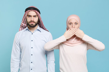 Shocked young couple friends arabian muslim man wonam in keffiyeh kafiya ring igal agal hijab clothes isolated on blue background studio. People religious lifestyle concept. Covering mouth with hands.