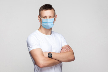 Portrait of funny young man in white shirt with surgical medical mask standing with crossed arms and looking at camera winking with funny face. indoor studio shot, isolated on gray background.
