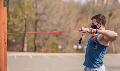 A young and athletic guy in a medical mask does exercises with sports elastic during a pandemic. COVID-19. Health care.