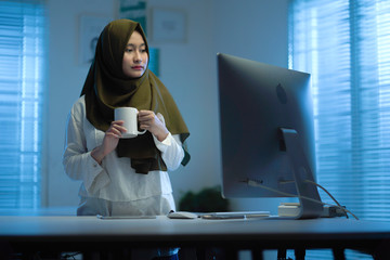 Beautiful Asian Muslims wearing darkgreen headscarves drinking coffee when working from home in a...