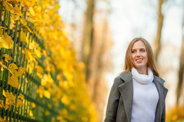 Beautiful girl with light brown hair walks along the alley of the autumn park. Attractive young woman in a gray coat and white sweater near the fence. Fall season. Yellow foliage. Amazing autumn mood.