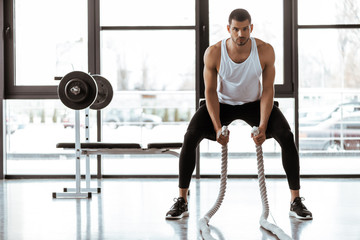 Naklejka na ściany i meble Handsome sportsman exercising with battle ropes in gym