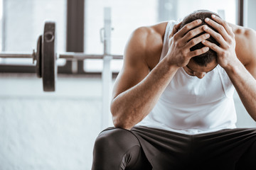 upset sportsman touching head while sitting near barbell in gym