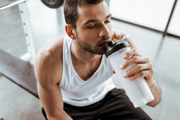 overhead view of sportsman drinking protein milkshake while holding sports bottle in gym