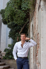 handsome and young guy in a white shirt enjoy the summer weather leans on a brick wall in Venice. © Andrii