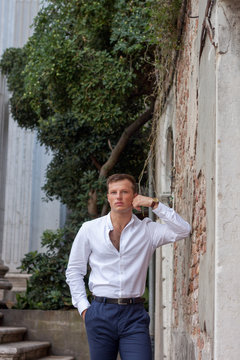 handsome and young guy in a white shirt enjoy the summer weather leans on a brick wall in Venice.