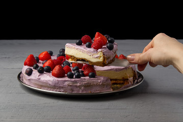 Woman divided cheesecake and take one slice to offer it. Home made Cheesecake with Strawberry, blueberry and raspberry on grey wooden background.
