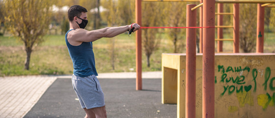 A young and athletic guy in a medical mask does exercises with sports elastic during a pandemic....