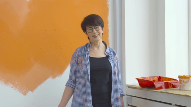 Flat renovation concept. Happy Middle-aged woman painting white wall with paint roller, orange paint