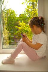child with a phone on the windowsill looks out the window. carontin. self-isolation. child at home. window. Street