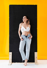 Beautiful busty girl wearing unbuttoned jeans and t-shirt poses at the black shield near yellow background. Fashionable, advertising, lifestyle and commercial design.