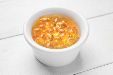 Bowl of fish oil capsules on white wooden background