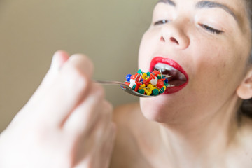 Beautiful girl with red lipstick eating colored sharp drawing pins from a metallic spoon. Conceptual image