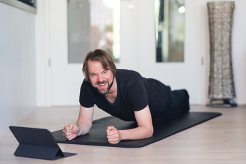 Mann macht Planks zu Hause auf der Fitnessmatte mittels Online-Kurs und lächelt in die Kamera