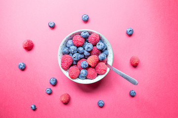 Close up of a green and white pot with spoon contining fresh 
raspberries and blueberries. Concept of healthy and seasonal food. Light red background