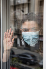 Girl with mask looking
through the window in a city apartment. Hand on the glass. City landscape in the shadow