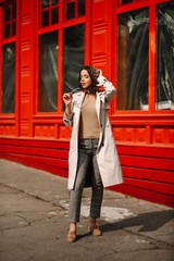 
Street fashion portrait of a young elegant luxurious lady in a beige trench coat, fashion sunglasses, shawl, posing on the street of a European city, against the background of a red building