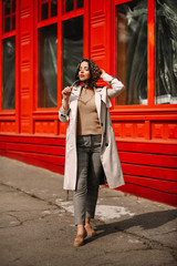 Obraz premium Street fashion portrait of a young elegant luxurious lady in a beige trench coat, fashion sunglasses, shawl, posing on the street of a European city, against the background of a red building